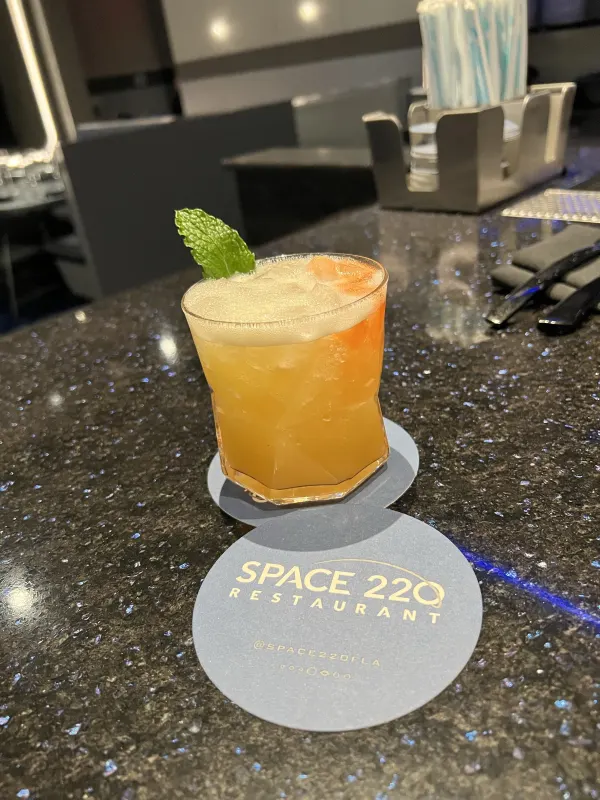 A signature cocktail on the branded coaster — the lounge prix fixe pairs well here.